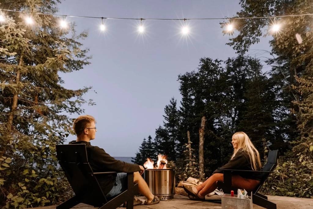 Top 5 Outdoor Amenities For HOAs & Condos 2 A man in a black jumper and a blonde girl in a black hoodie sit by an outdoor firepit under string lights