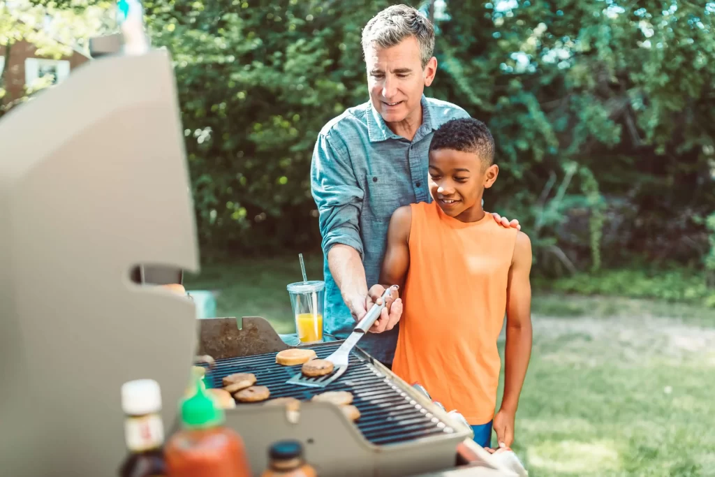 Top 5 Outdoor Amenities For HOAs & Condos 3 older white man in blue shirt shows young black boy in an orange top how to grill a burger. HOA outdoor amenities.