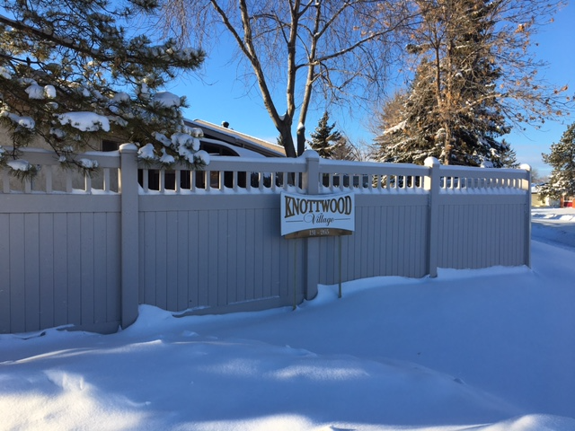 Snowy light brown fence with a sign that says Knottwood Village. There is snow piled up in front and with trees behind